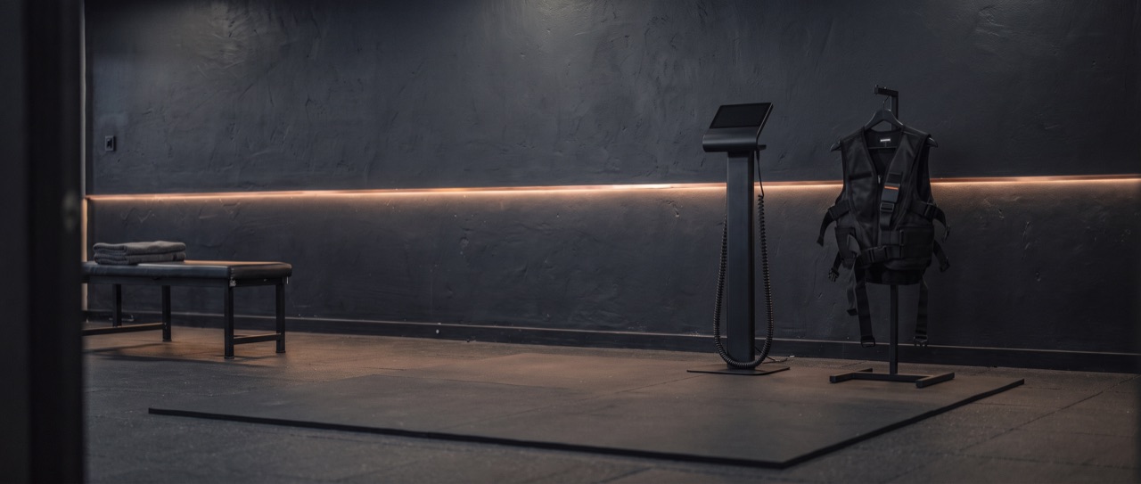 One training station in the corner of a room that carries on past it: a slim control unit, a training vest on a stand, and a mat laid on the floor